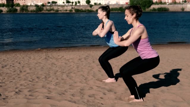 Two-woman-doing-yoga-on-the-beach-by-the-river-in-the-city.-Beautiful-view.-Utkatasana-pose.