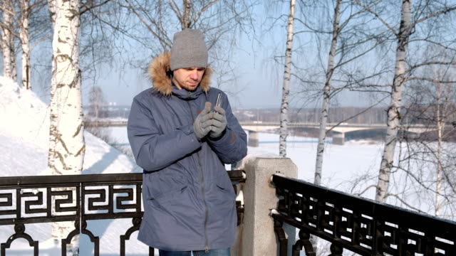 Mann-in-blau-Daunenjacke-mit-Fell-Kapuze-nehmen-sein-Handy-und-zieht-den-Handschuh-in-einem-Winter-Park.