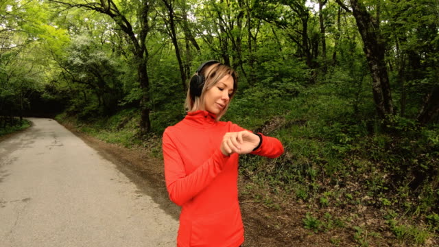 Young-attractive-woman-in-headphones-changing-the-settings-on-a-smart-watch-in-front-of-or-psole-jogging-on-the-road-in-a-green-forest