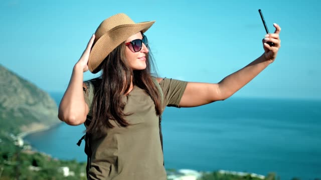 Portrait-of-smiling-woman-traveler-in-hat-and-sunglasses-posing-and-taking-selfie-using-smartphone