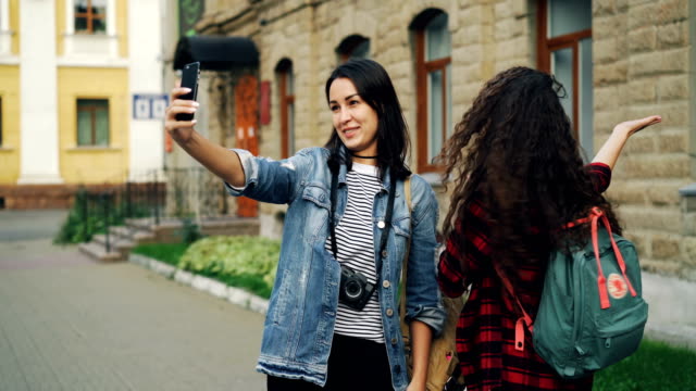 Emotionale-junge-Frauen-machen-Reisende-online-video-Anruf-mit-Smartphone-Haltevorrichtung-und-zeigt-historische-Gebäude-hinter-ihnen-im-Gespräch-mit-dem-Ausdruck-ihrer-Aufregung.