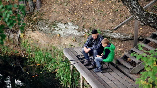 Little-boy-is-fishing-in-lake-with-his-daddy-from-wooden-pier-on-autumn-day,-child-is-holding-rod-sitting-on-chair-while-his-father-is-talking-to-him.-Nature-and-people-concept.