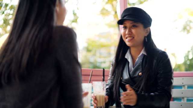 Gerne-Asiatin-ist-im-Gespräch-mit-ihrer-Freundin-im-Straßencafé-dann-Toasten-und-Klirren-der-Gläser-mit-Cocktails-genießen-treffen.-Kommunikation-und-Getränke-Konzept.