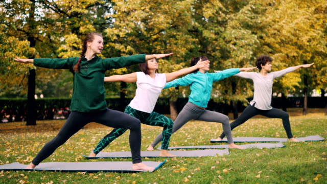 Señoritas-estudiantes-de-yoga-están-de-pie-en-actitud-de-Guerrero-y-pasando-posteriormente-a-su-posición-de-triángulo-durante-la-clase-al-aire-libre-en-el-parque-en-otoño.-Concepto-de-juventud-y-salud.