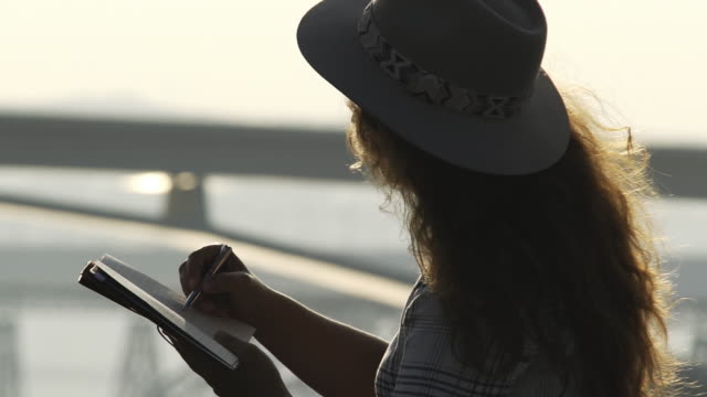 Woman-writing-in-notebook-and-looking-at-sunrise-on-bridge