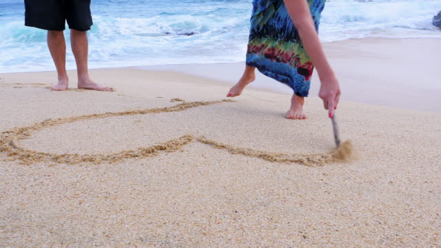 An-older-woman-draws-a-heart-and-initials-in-the-sand-and-her-husband-gives-her-a-kiss
