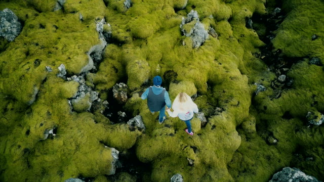 Top-aerial-view-of-the-young-beautiful-couple-walking-on-the-lava-field-covered-moss-in-Iceland,-exploring-the-nature