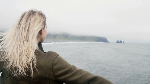 Rückansicht-der-jungen-blonden-Frau-am-schwarzen-Strand-in-Island.-Freiheit:-touristische-erhebt-sich-Hände,-Haar-Welle-auf-wind