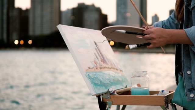 Mujer-joven-de-dibujo-en-la-orilla-del-lago-Michigan-en-Chicago,-América.-Luces-de-la-ciudad-en-el-fondo