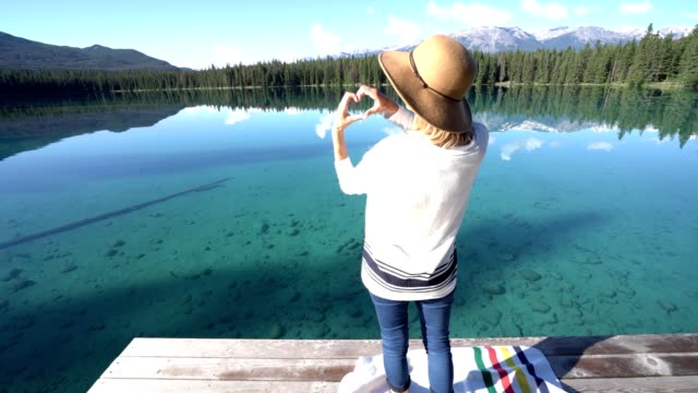 Young-woman-on-wooden-pier-makes-heart-shape-frame-in-nature