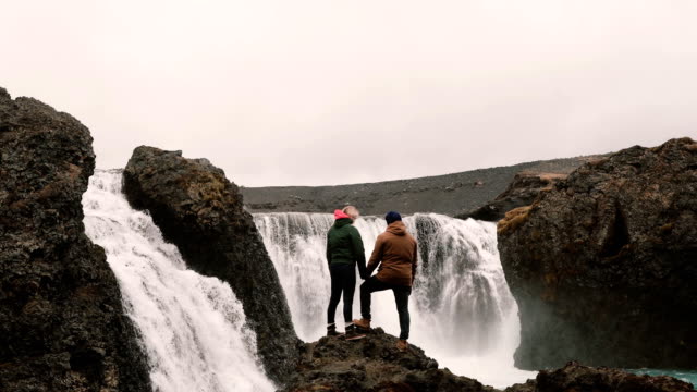 Vista-posterior-de-una-pareja-joven-de-pie-sobre-una-roca-y-mirando-en-una-potente-cascada-en-Islandia,-las-manos