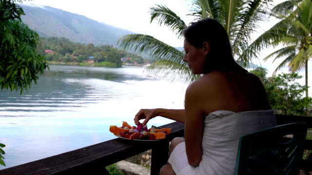 Eine-Frau-sitzt-in-einem-Handtuch-vor-dem-Hintergrund-von-einem-See-und-Berge-und-frisst-die-Pitahaya