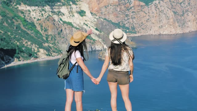 Back-view-two-active-female-tourists-smiling-enjoying-magnificent-view-of-sea-and-mountain