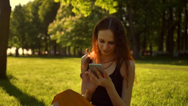 Happy-Smiling-Young-Woman-enjoying-Nature-and-sunrise.-Sitting-on-bench-in-green-summer-park-using-mobile