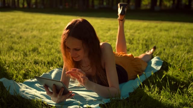 Female-sitting-in-the-park-among-the-trees-and-use-the-Internet-on-tablet