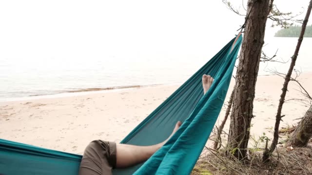Young-caucasian-male-resting-lying-on-blue-hammock-outdoors-near-lake.