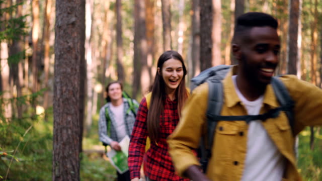 Cheerful-African-American-man-is-holding-map-and-asking-his-friends-to-follow-him-during-trip-in-forest-on-warm-summer-day.-Hiking,-nature-and-people-concept.