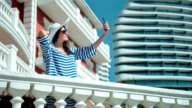Glamour-Frau-in-Hut-und-eine-Sonnenbrille-nehmen-Selfie-mit-Smartphone-stehen-auf-der-Hotelterrasse