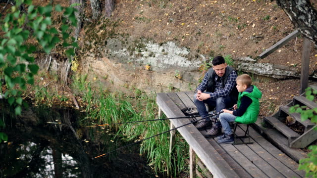 High-angle-view-of-fisherman-and-his-child-serious-boy-fishing-from-wooden-pier-sitting-on-chairs-with-rods-and-talking.-Family,-hobby,-nature-and-relationship-concept.