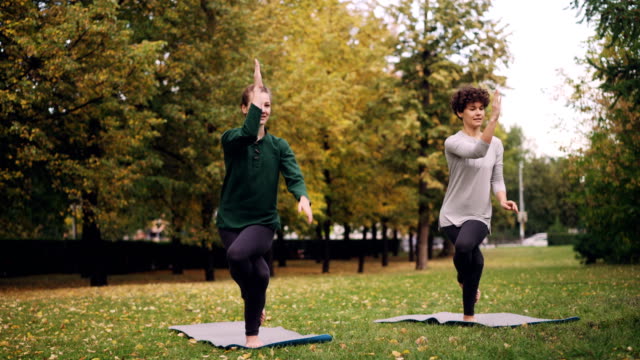 Slender-girl-yoga-student-is-learning-Eagle-pose-under-guidance-of-teacher-during-individual-practice-with-instructor-in-park.-Beautiful-autumn-nature-is-in-background.