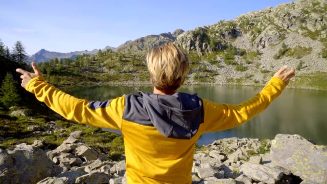Joven-excursionista-en-ruta-en-verano-por-el-impresionante-lago-alpino-brazos-extendidos-celebrando-personal-objetivo.-Hombre-joven-de-pie-en-la-naturaleza-amplia-de-brazos-abiertos