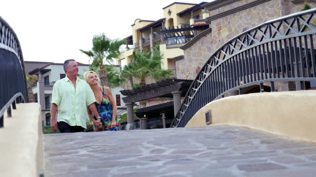 Eine-ältere-paar-Hände-halten,-zu-Fuß-über-einer-kleinen-Brücke-in-einem-Resort