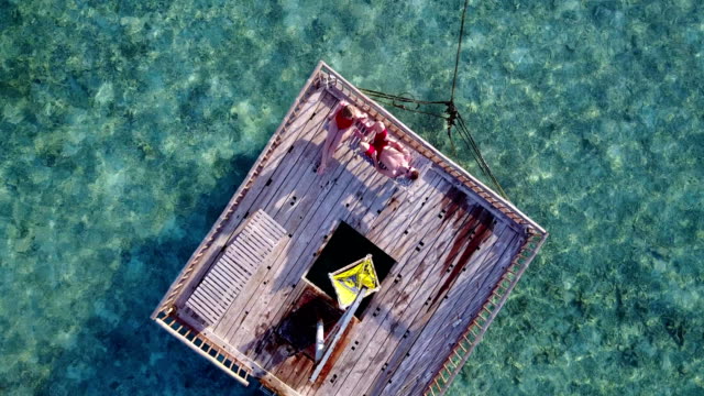 v03824-Aerial-flying-drone-view-of-Maldives-white-sandy-beach-2-people-young-couple-man-woman-relaxing-on-sunny-tropical-paradise-island-with-aqua-blue-sky-sea-water-ocean-4k-floating-pontoon-jetty-sunbathing-together