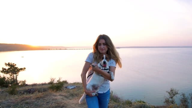Young-attractive-woman-playing-with-a-dog-Jack-Russell-in-the-meadow-at-sunset-with-sea-background.-slow-motion