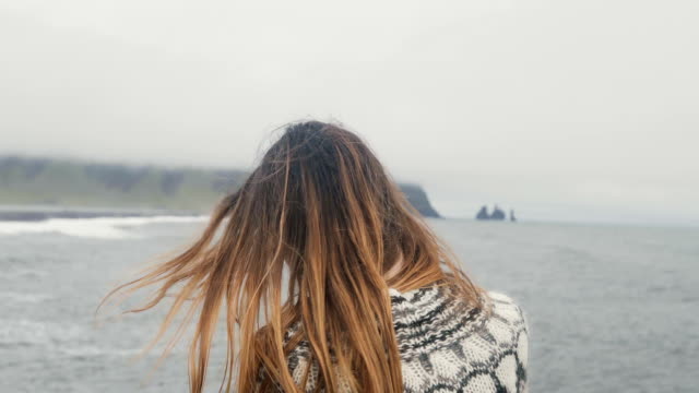 Back-view-of-young-brunette-woman-standing-on-the-black-beach-and-enjoying-beautiful-view-of-troll-toes-rocks-in-Iceland