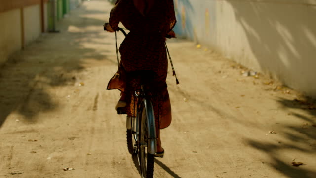 Following-Shot-of-a-Beautiful-Woman-Wearing-Dress-Rides-Bicycle-Through-Narrows-Streets-of-Small-Town.-South-Asia-Nature,-Palm-Trees-Grow-by-the-Road.-Slow-Motion.