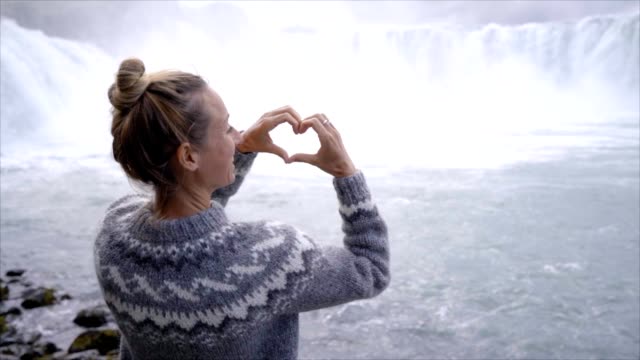 LANGSAME-Bewegung-der-jungen-Frau-in-Island-macht-Herz-Form-Finger-Frame-auf-spektakulären-Wasserfall