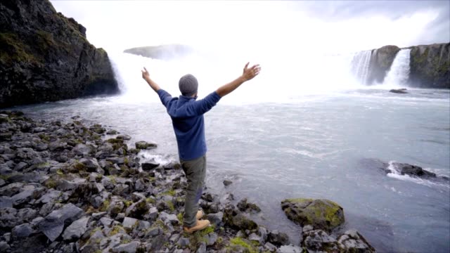 Junger-Mann,-Arme-ausgestreckt-vor-dem-herrlichen-Wasserfall-in-Island