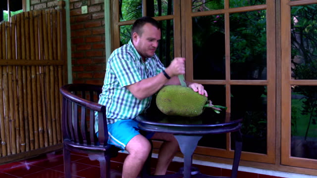 Ein-Mann-sitzt-auf-der-Terrasse-die-Jackfruit-mit-einem-Messer-Aufschlitzen