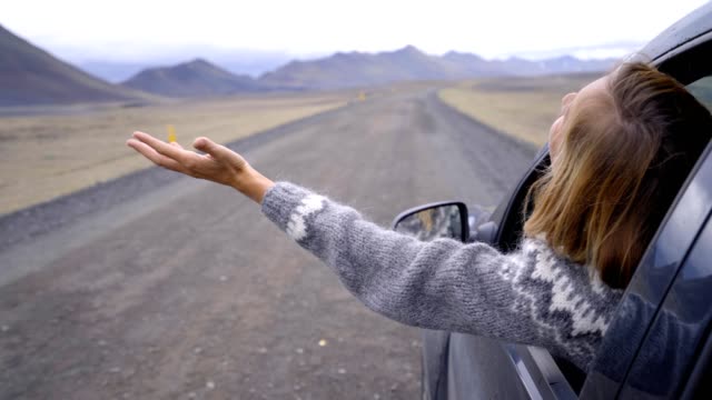 Cheerful-caucasian-female-inside-car-looking-at-road-arms-outstretched-,-mountain-volcanic-landscape.-Road-trip-concept.--Blond-hair-girl-looking-at-landscape.