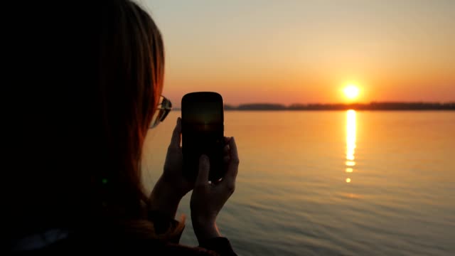 Mädchen-übernimmt-Bilder-von-traumhaften-Sonnenuntergang-und-Fluss-Böschung-durch-ihr-modernes-Mobiltelefon
