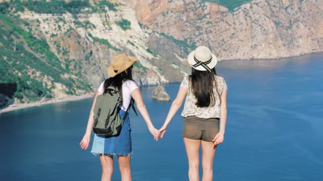 Two-active-elegant-young-traveler-raising-hands-and-having-fun-standing-on-cliff-edge-rear-view