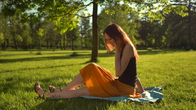 Happy-smiling-attractive-woman-sitting-on-green-grass-and-have-conversation-via-digital-smartphone-device