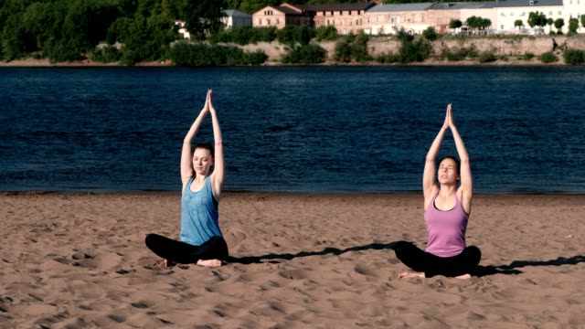 Two-woman-stretching-yoga-on-the-beach-by-the-river-in-the-city.-Beautiful-city-view.-Namaste-pose.