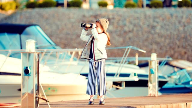 Small-and-beautiful-girl-looks-through-binoculars-at-the-wharf-for-boats-and-yachts