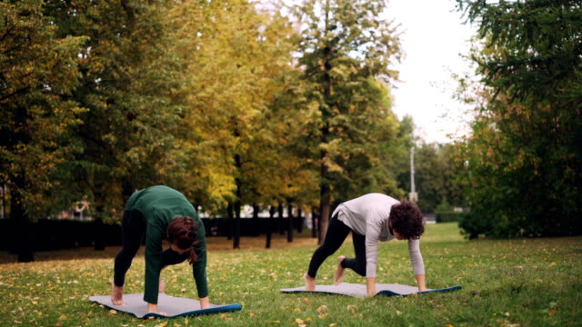 Smiling-girls-yoga-teacher-and-student-are-exercising-outdoors-on-grass-in-park-practising-asanas-on-mats-with-green-and-yellow-trees-around.-Healthcare-and-sports-concept.
