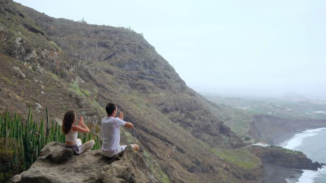 Un-hombre-y-una-mujer-sentada-en-la-cima-de-una-montaña-mirando-el-mar-sentado-en-una-piedra-meditando-en-posición-de-loto.-La-vista-desde-la-parte-posterior.-Islas-Canarias