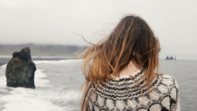 Young-brunette-woman-standing-on-the-shore-of-the-sea-and-looking-on-troll-toes-sight-in-Iceland,-hair-waving-on-wind