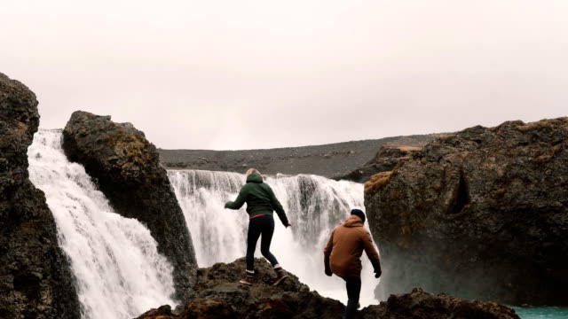 Young-happy-couple-hiking-together-to-see-a-powerful-waterfall-in-Iceland.-Man-and-woman-raising-hands-up