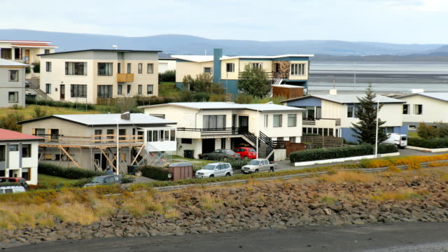 Bau--und-geparkte-Autos-in-Außenbezirken-von-isländischen-Kleinstadt,-Fluss-und-die-Berge-sind-im-Hintergrund