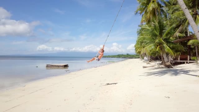 Drone-shot-aerial-view-of-young-man-playing-on-beach,-swing-rope-on-palm-tree.-Shot-in-the-Philippines,-4K-resolution-video.-People-travel-vacations-concept