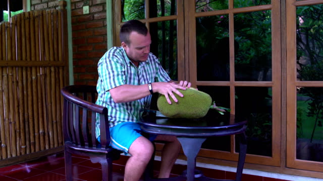 Ein-Mann-sitzt-auf-der-Terrasse-und-die-Jackfruit-mit-einem-Messer-schneidet