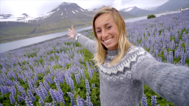 Selfie-portrait-of-tourist-female-in-Iceland-in-the-middle-of-Lupine-purple-flowers,-smiling-hair-in-wind,-wool-seater