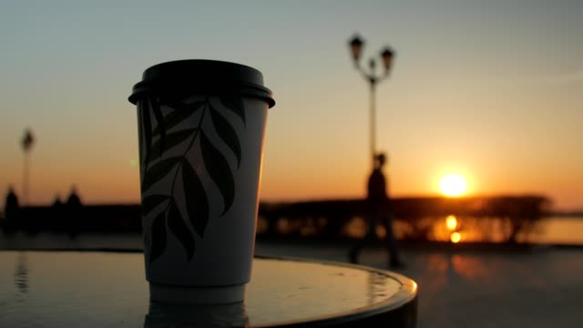 una-taza-de-café-está-de-pie-sobre-una-mesa-de-un-café-de-calle-en-el-fondo-del-terraplén,-puesta-de-sol-cielo
