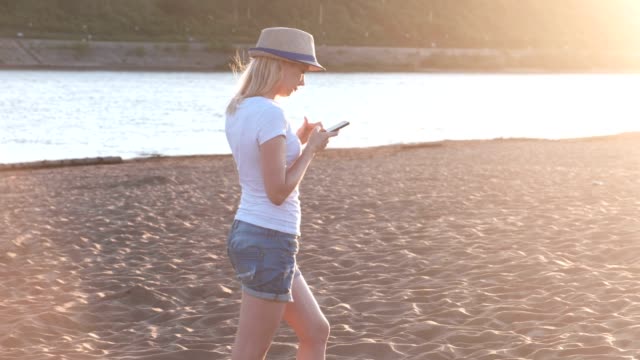 Chica-rubia-hermosa-con-un-sombrero-tipo-un-mensaje-en-su-teléfono-móvil-caminando-por-la-playa-al-atardecer.