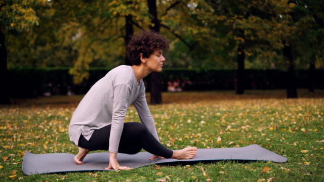 Slim-young-lady-is-doing-sequence-of-yoga-asanas-sitting-on-mat-in-park-alone-on-autumn-day-with-beautiful-grass-and-trees-around-her.-Recreation-and-sport-concept.